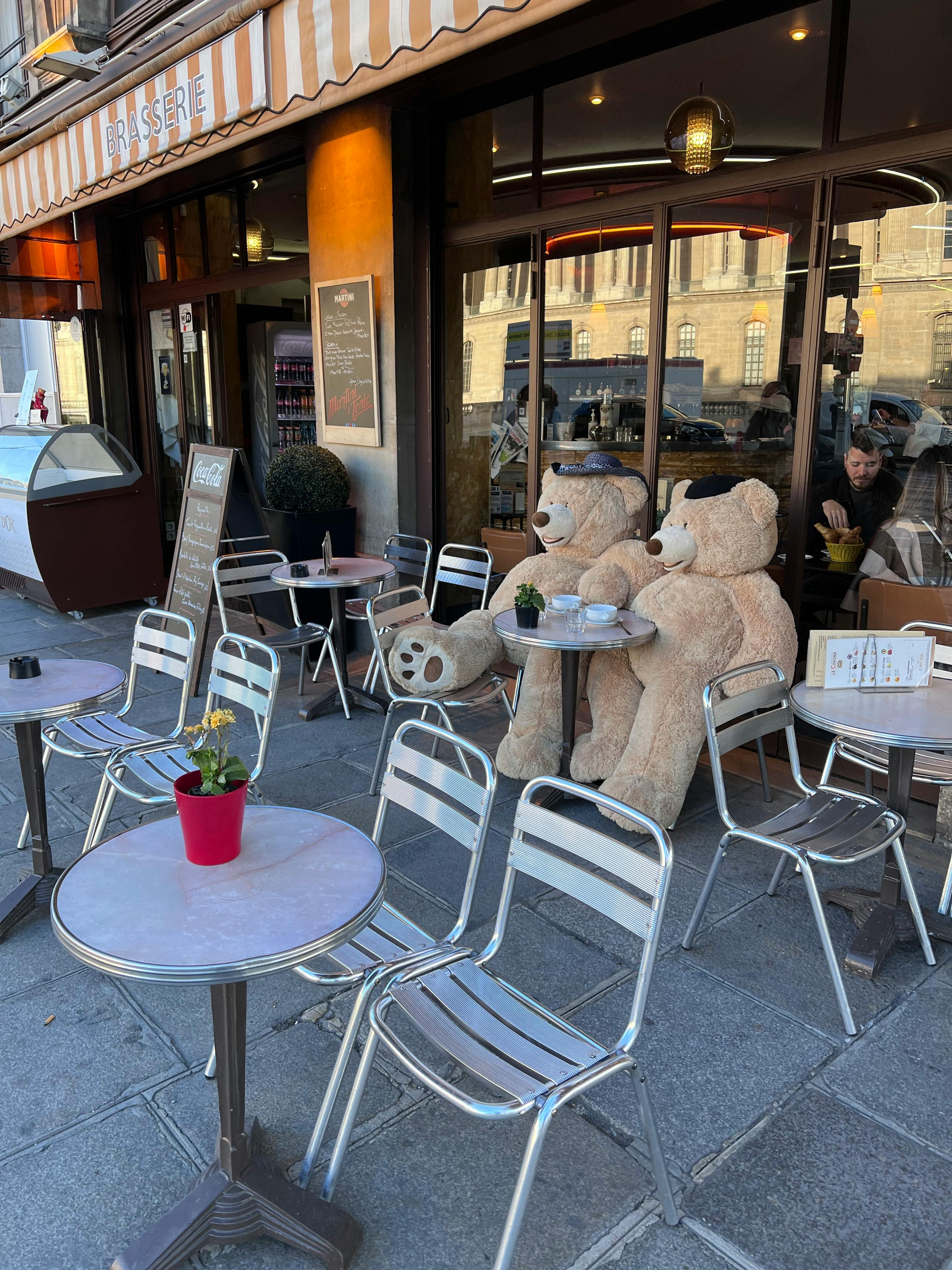 Outdoor café featuring large teddy bears seated at metal tables with a street view.