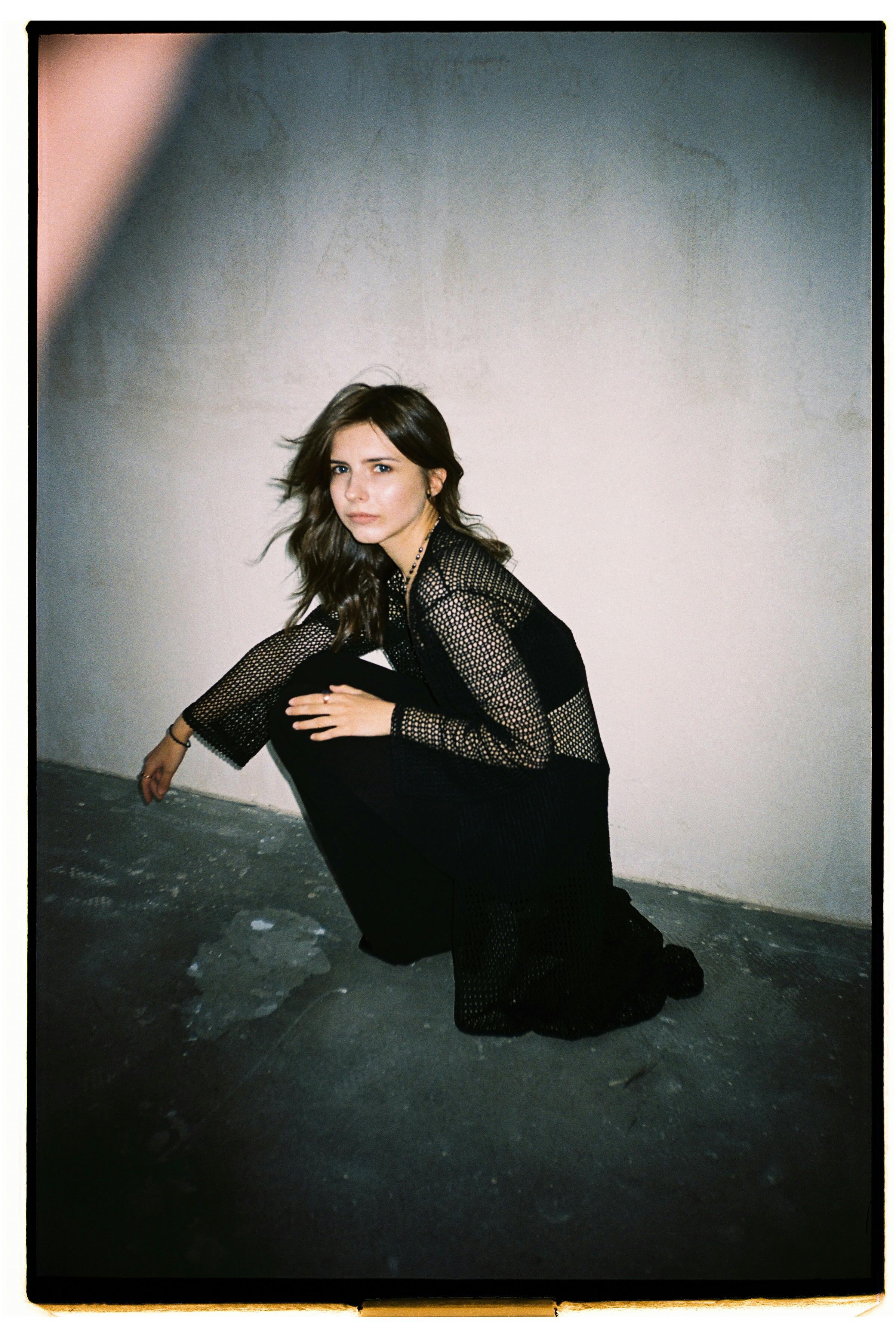 A well-dressed woman crouches on a gritty indoor floor, exuding mystery and elegance.