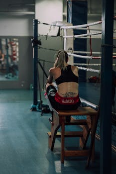 Female boxer in a sports bra resting on a bench in a boxing gym environment.