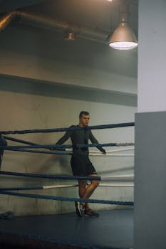 A male boxer resting against ropes in an indoor boxing ring, focused and relaxed.