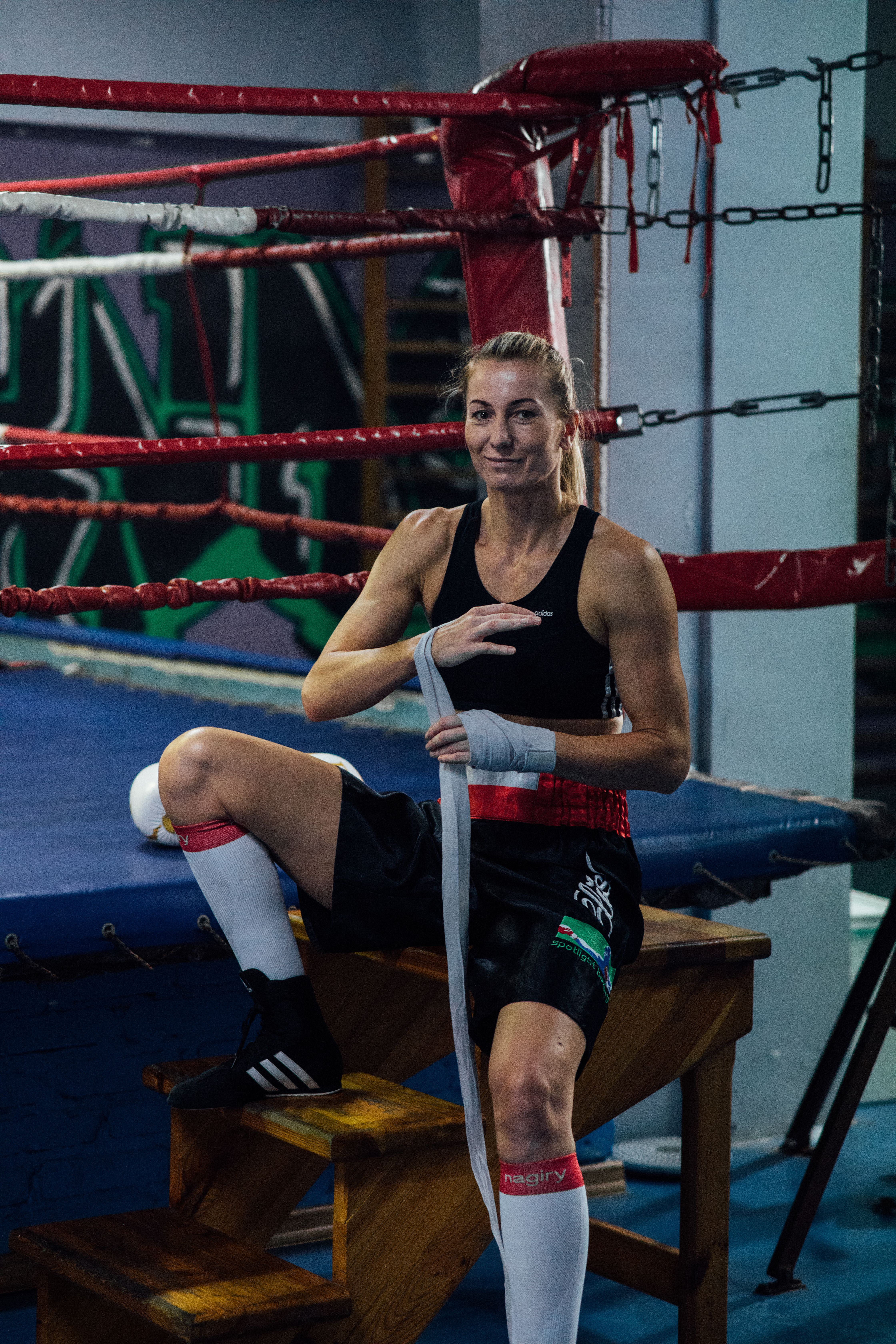 A female boxer sitting in a gym, wrapping her hands in front of a boxing ring, smiling confidently.