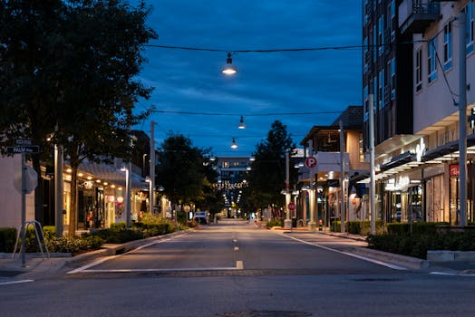 Empty street scene with storefronts and street lights at twilight in an urban setting.
