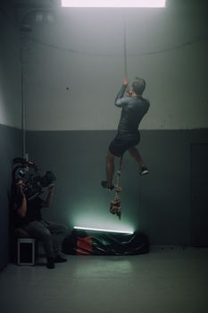 A strong man climbing a rope in an indoor gym with dim lighting, showcasing athletic strength.