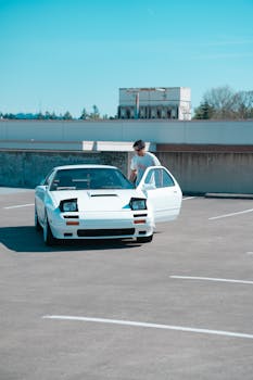 Man getting into a classic white sports car in an empty parking lot under a clear blue sky.