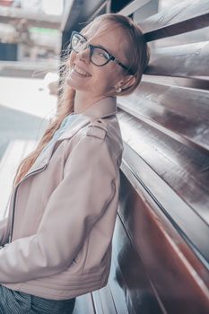 Caucasian woman smiling while leaning against a wooden wall, wearing glasses and a stylish jacket.