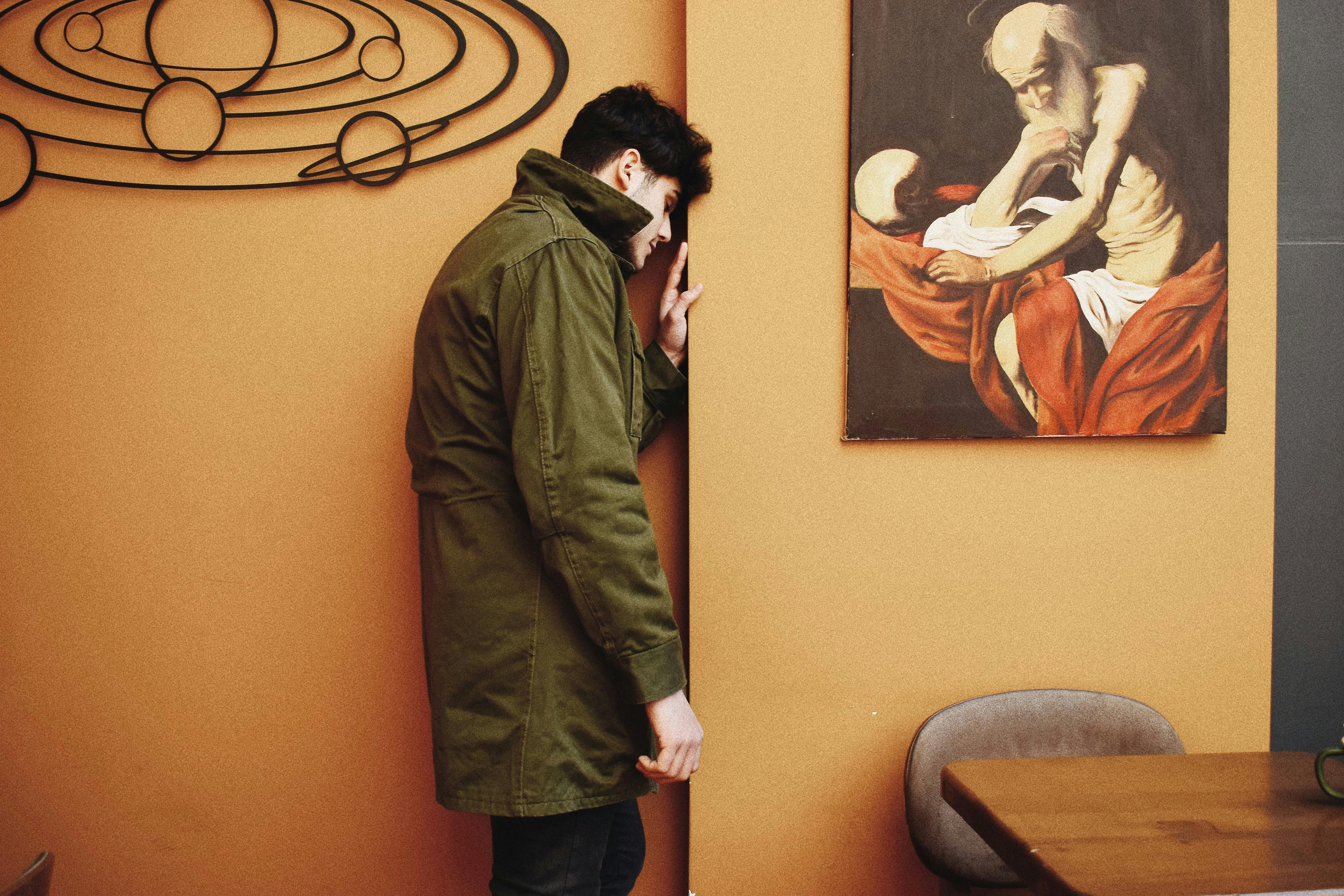 A man leans on a wall indoors, next to a painting and décor.