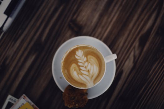 A cappuccino with intricate latte art in a white cup, placed on a wooden table, viewed from above.