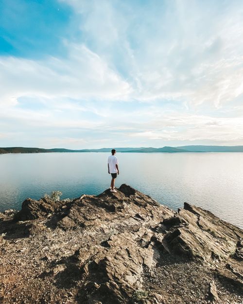 Free A lone man stands on a rocky formation, gazing over a serene lake under a vast sky. Stock Photo