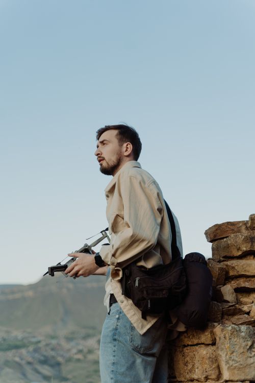 Free Man standing by stone wall holding drone, with scenic mountain backdrop. Stock Photo