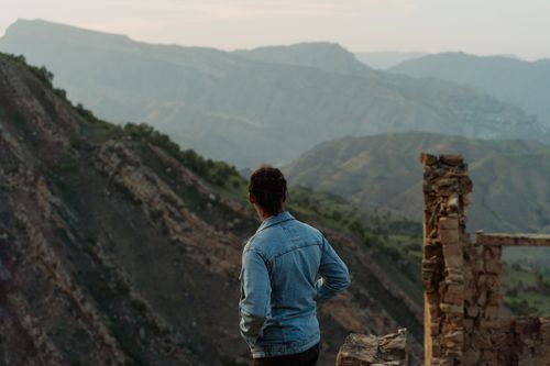 Free A man wearing a denim jacket stands and gazes at a vast mountain range during the day. Stock Photo