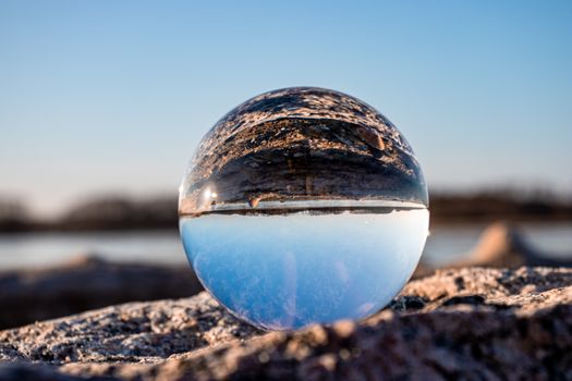A reflective glass ball capturing an inverted landscape on a rocky beach under a clear blue sky.