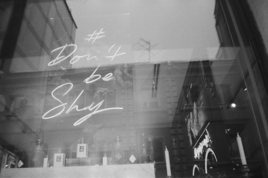 Black and white photo of a neon sign reading '# Don't be Shy' reflected in a window.