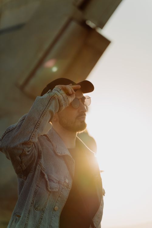 Free Young man wearing a denim jacket and sunglasses, standing outdoors in bright sunlight, exuding style and confidence. Stock Photo