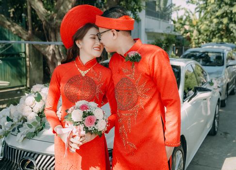 A smiling couple in red traditional attire standing beside a decorated car, outdoors.