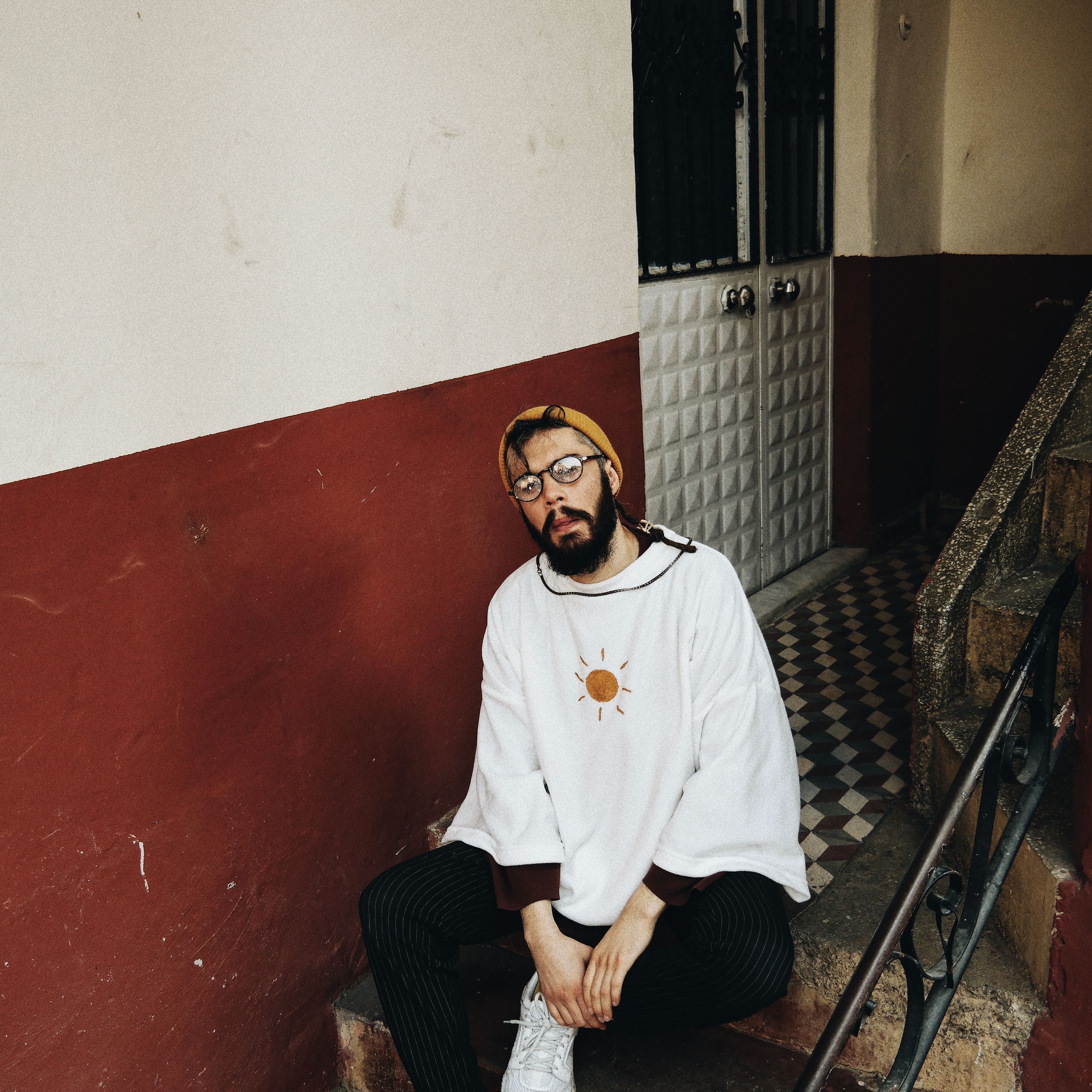 Fashionable young man with beard and glasses sitting on a staircase, showcasing urban style.