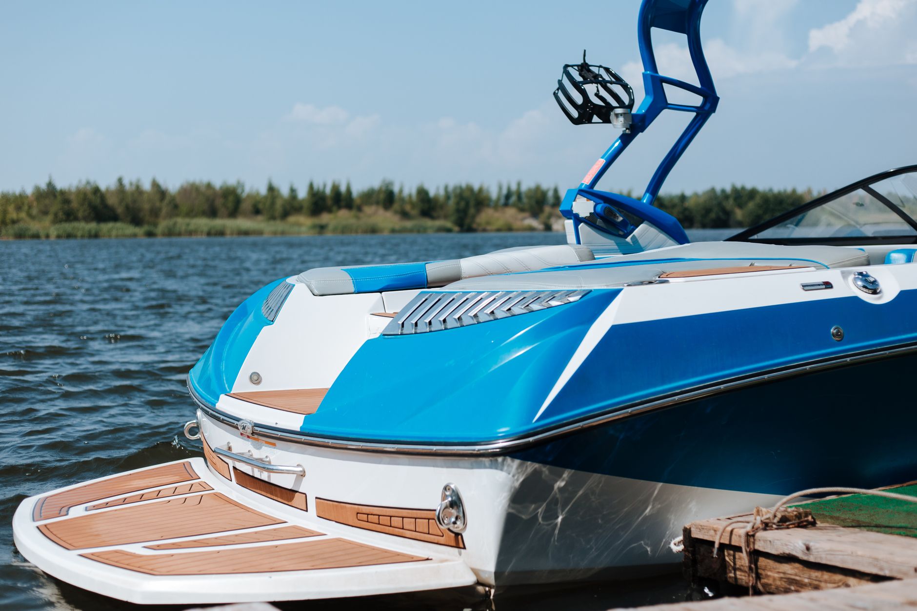 A sleek blue motorboat moored at a sunny lakeshore under a clear sky.
