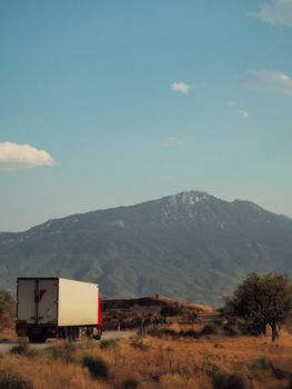 A truck navigates a rural road under a blue sky with mountains in the background.
