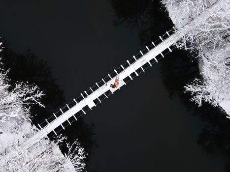 Drone shot of a couple lying on a snow-covered wooden bridge over a dark river in a winter landscape.