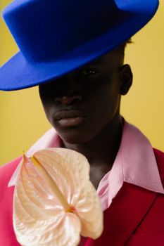 Chic studio portrait of a man in a blue hat holding a pink anthurium against a yellow background.