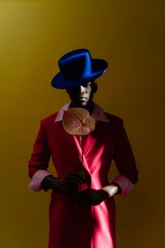 Fashionable man in vibrant pink coat and blue hat holding a flower against a yellow background.