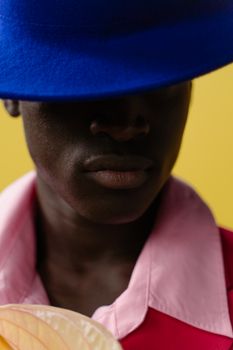 Close-up of a fashionable individual in a blue hat and pink shirt against a yellow studio backdrop.