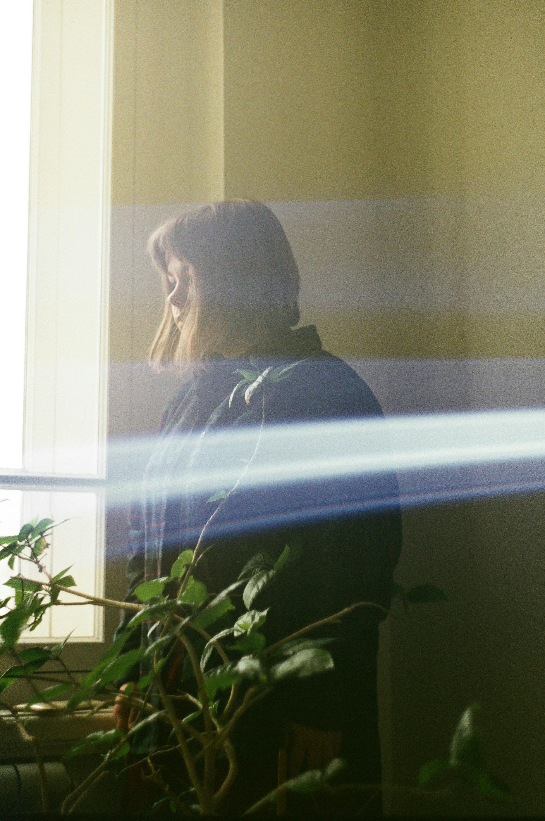 A woman stands by a window, partially obscured by streaks of light, in a serene indoor setting.