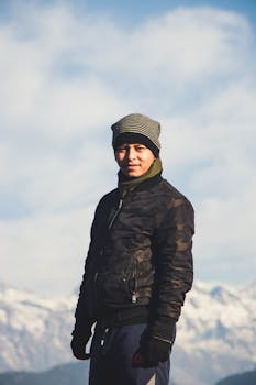 A man in winter clothes smiles against a snowy mountain backdrop, showcasing winter fashion.