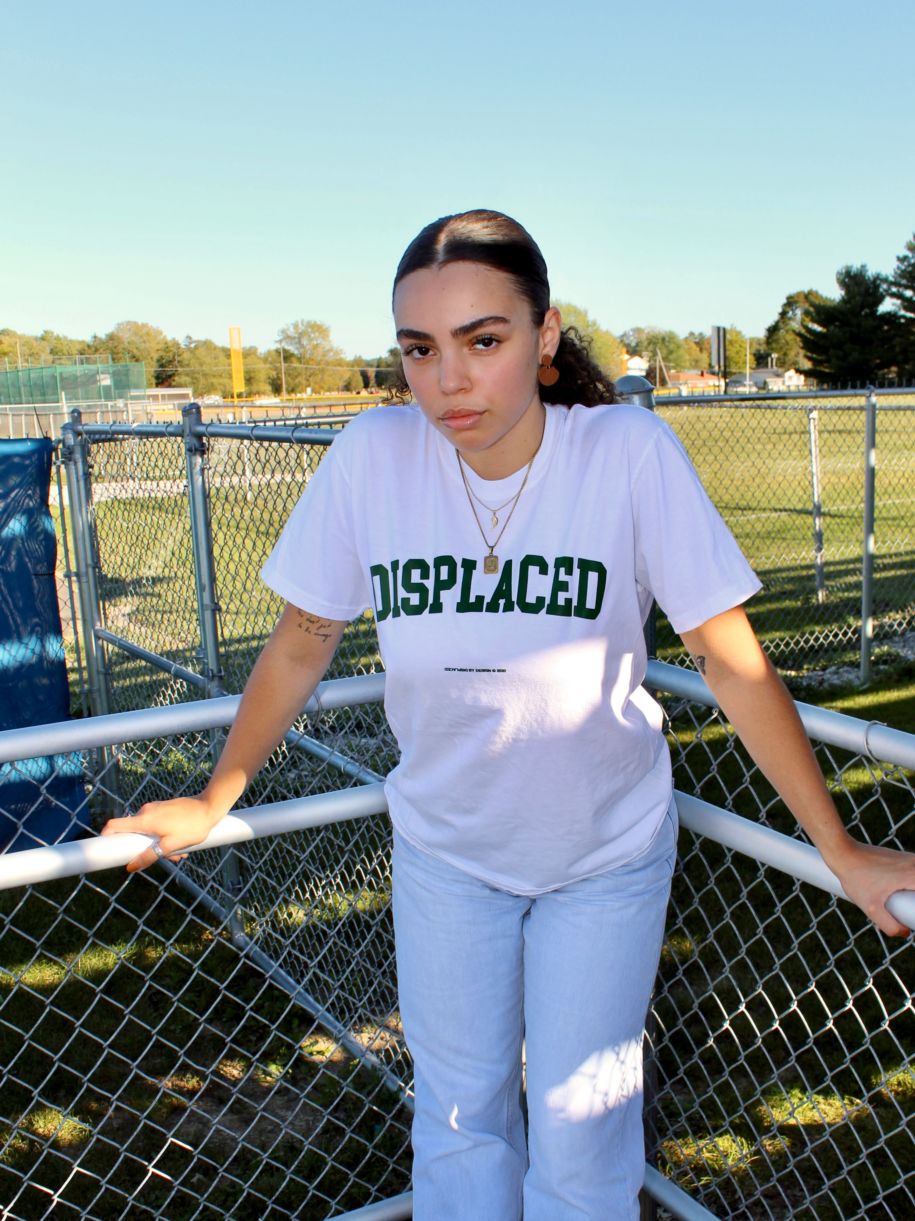 A young woman in a white 'DISPLACED' shirt leans on a fence in a park setting.