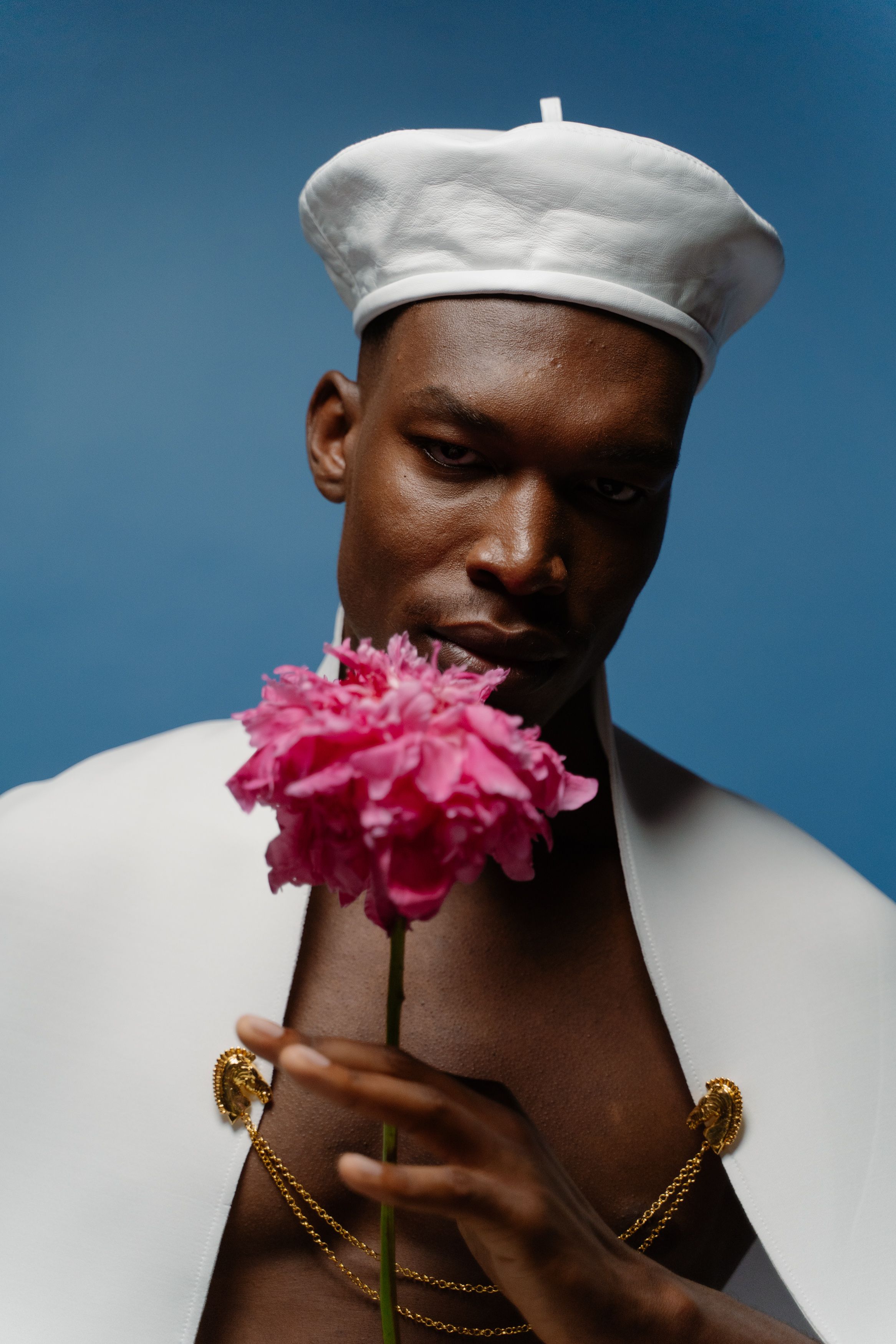 Stylish studio portrait of a man holding a pink flower, wearing a white beret and cape.