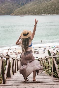 Woman wearing a sarong and summer hat poses on wooden stairs leading to a Brazilian beach.