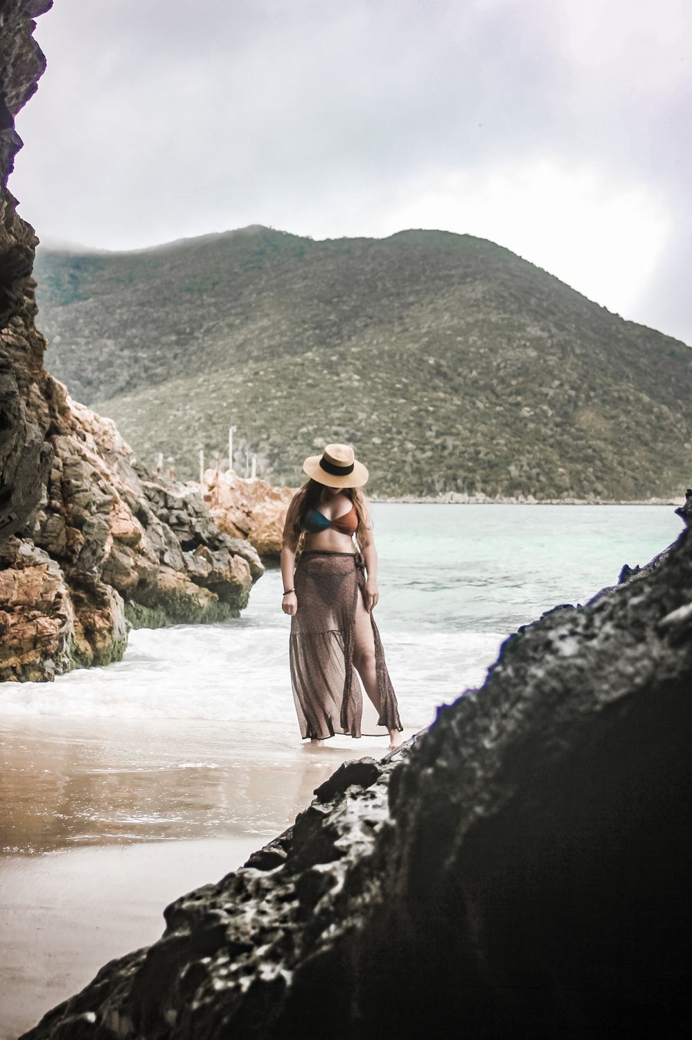 Woman in straw hat standing on a rocky tropical beach with mountains in the background