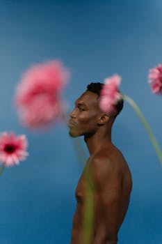 Artistic studio portrait of a shirtless black man posing with pink flowers against a blue background.