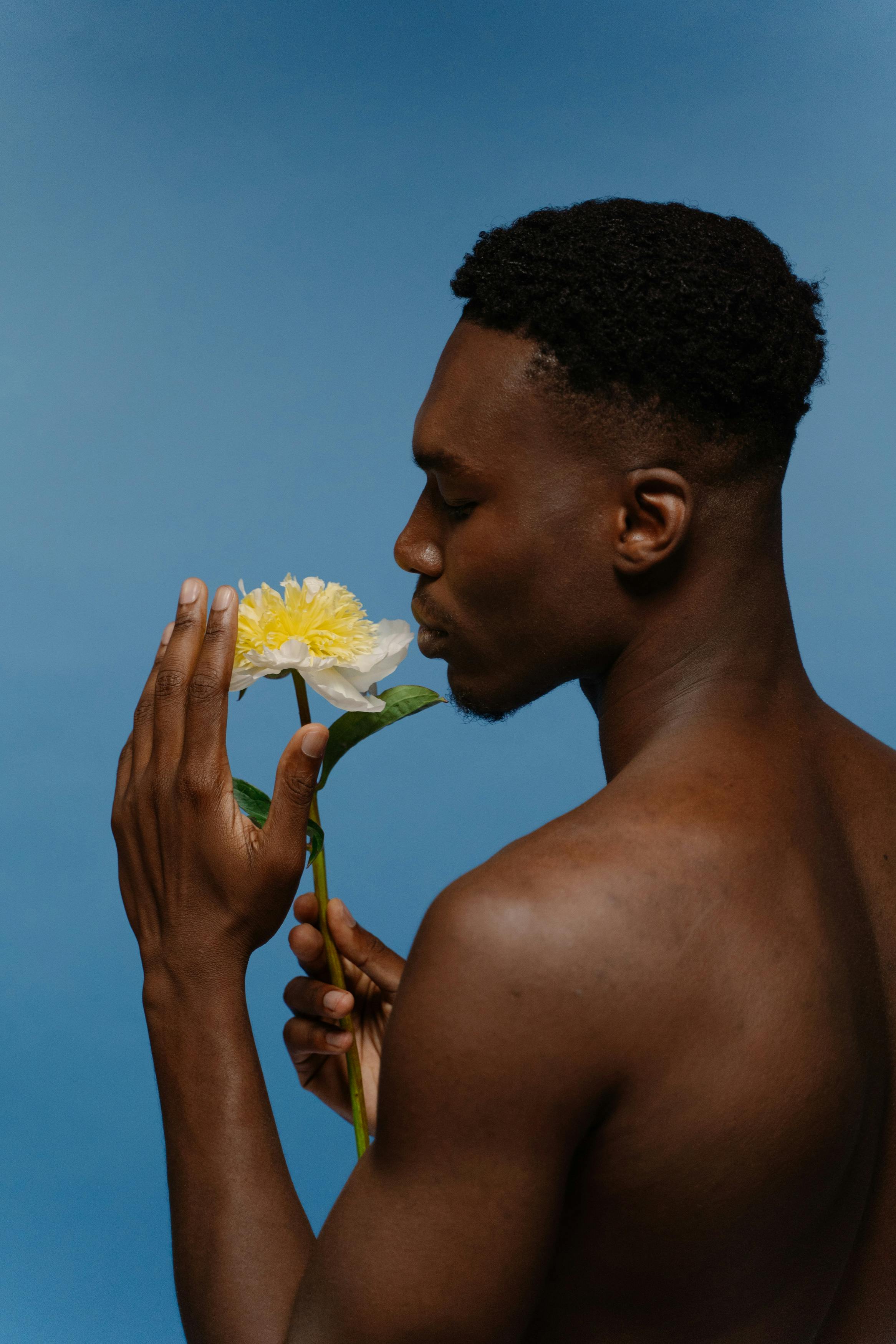 A serene studio portrait of a shirtless man smelling a yellow peony flower against a blue background.