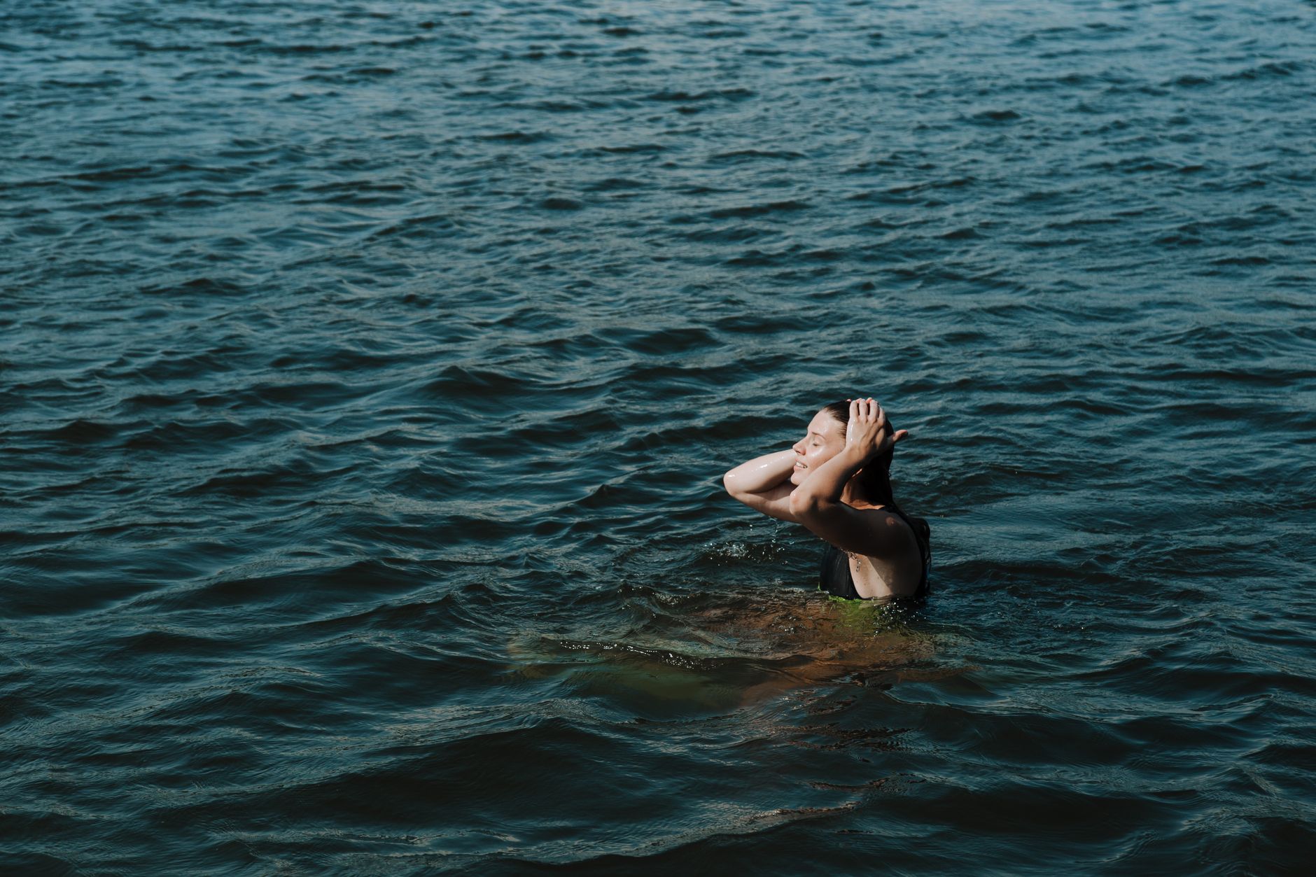 A woman with eyes closed enjoys a refreshing swim in a calm lake, touching her hair.