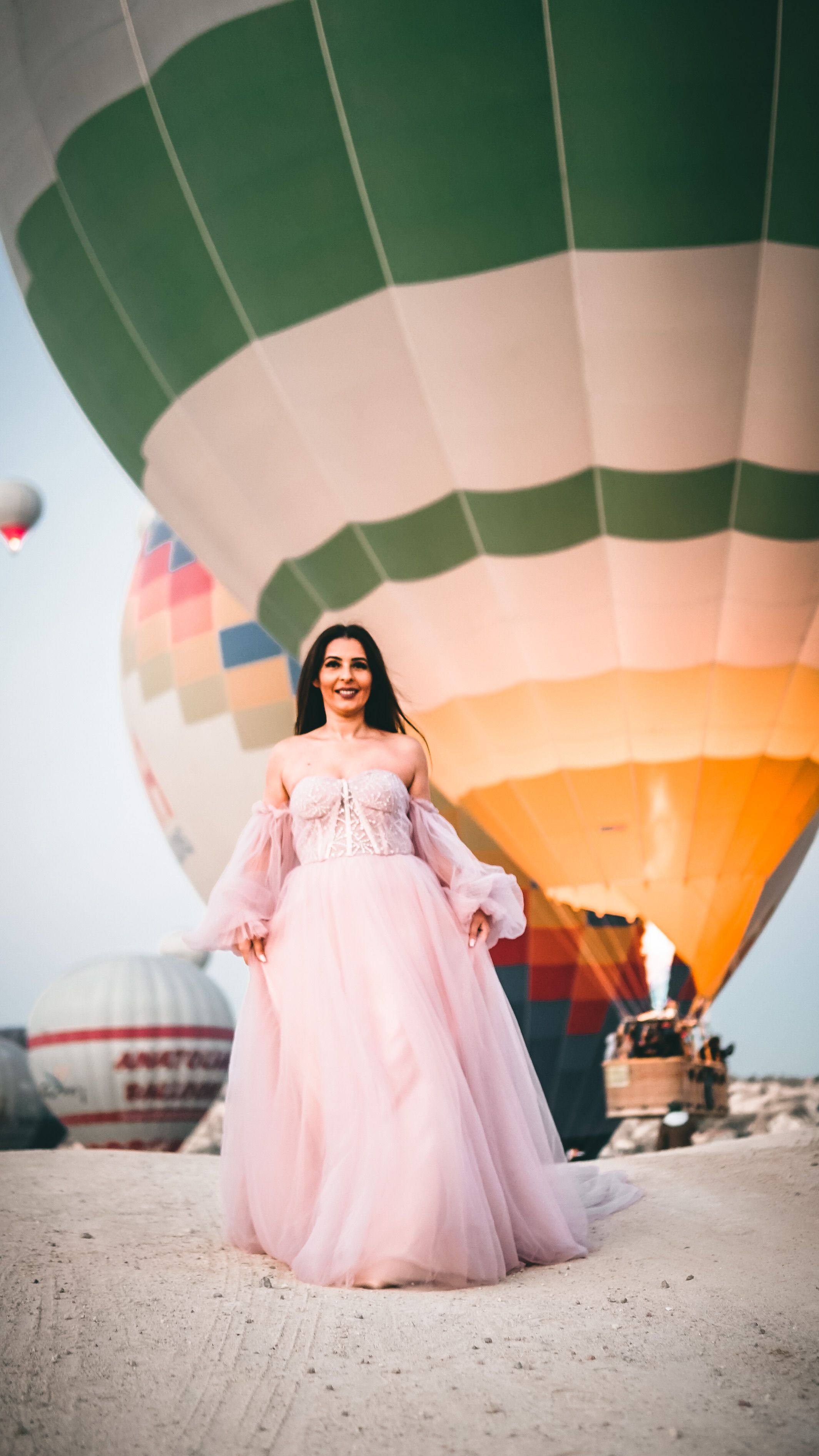 A happy woman in a pink dress stands gracefully in front of colorful hot air balloons during daytime.