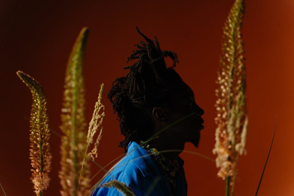 Artistic portrait of a stylish man with dreadlocks surrounded by plants against an orange studio backdrop.