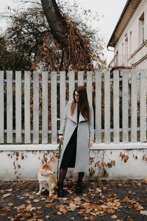 Woman in Gray Coat and Her Pet Dog Standing on Pavement with Fallen Leaves