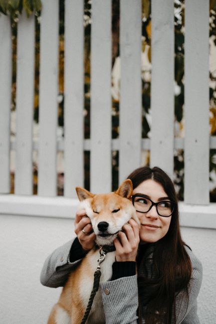 A cheerful woman with glasses holding her Shiba Inu dog outdoors by a fence.