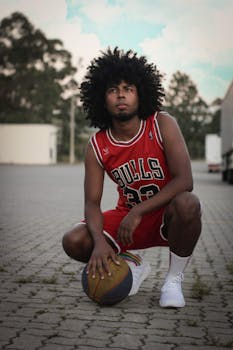 Young man with afro hair in basketball jersey crouching with ball on pavement.