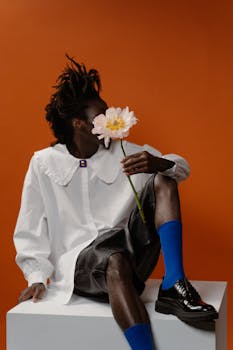Fashion portrait of a person in stylish attire holding a flower against orange backdrop.