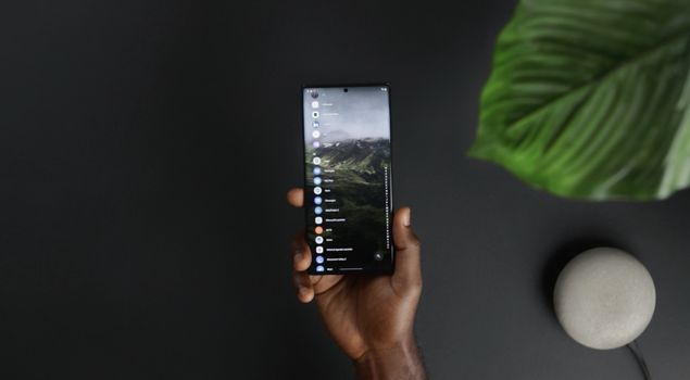 A hand holding a smartphone indoors with apps displayed on the screen.