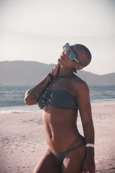Confident woman posing on a serene beach, showcasing tattoos and modern sunglasses under the sun.