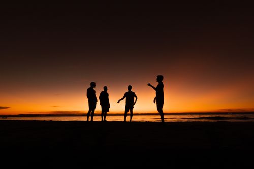 Free Silhouetted figures of four friends standing on a beach, expressing joy during a vibrant sunset. Stock Photo