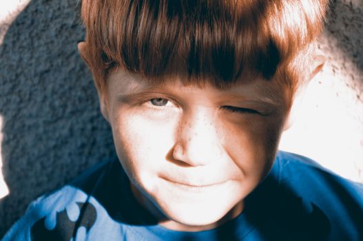 Close-up of a young freckled boy wearing a blue shirt, winking and smiling in bright sunlight.