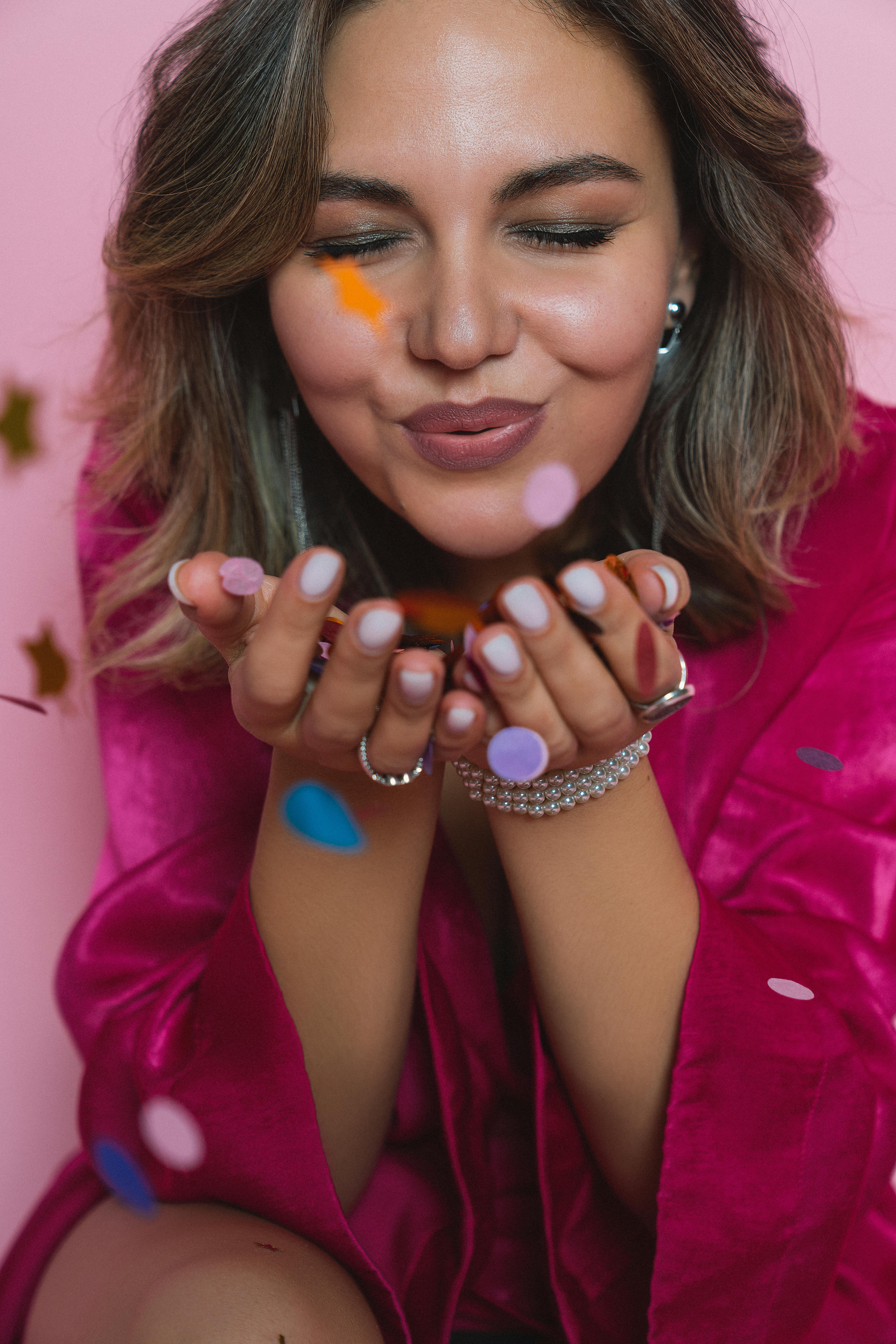 Joyful woman in a pink robe blowing confetti, smiling in a studio setting.