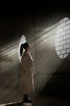 A woman stands in a sunlit warehouse, enveloped in dramatic light and shadows, wearing a trench coat.
