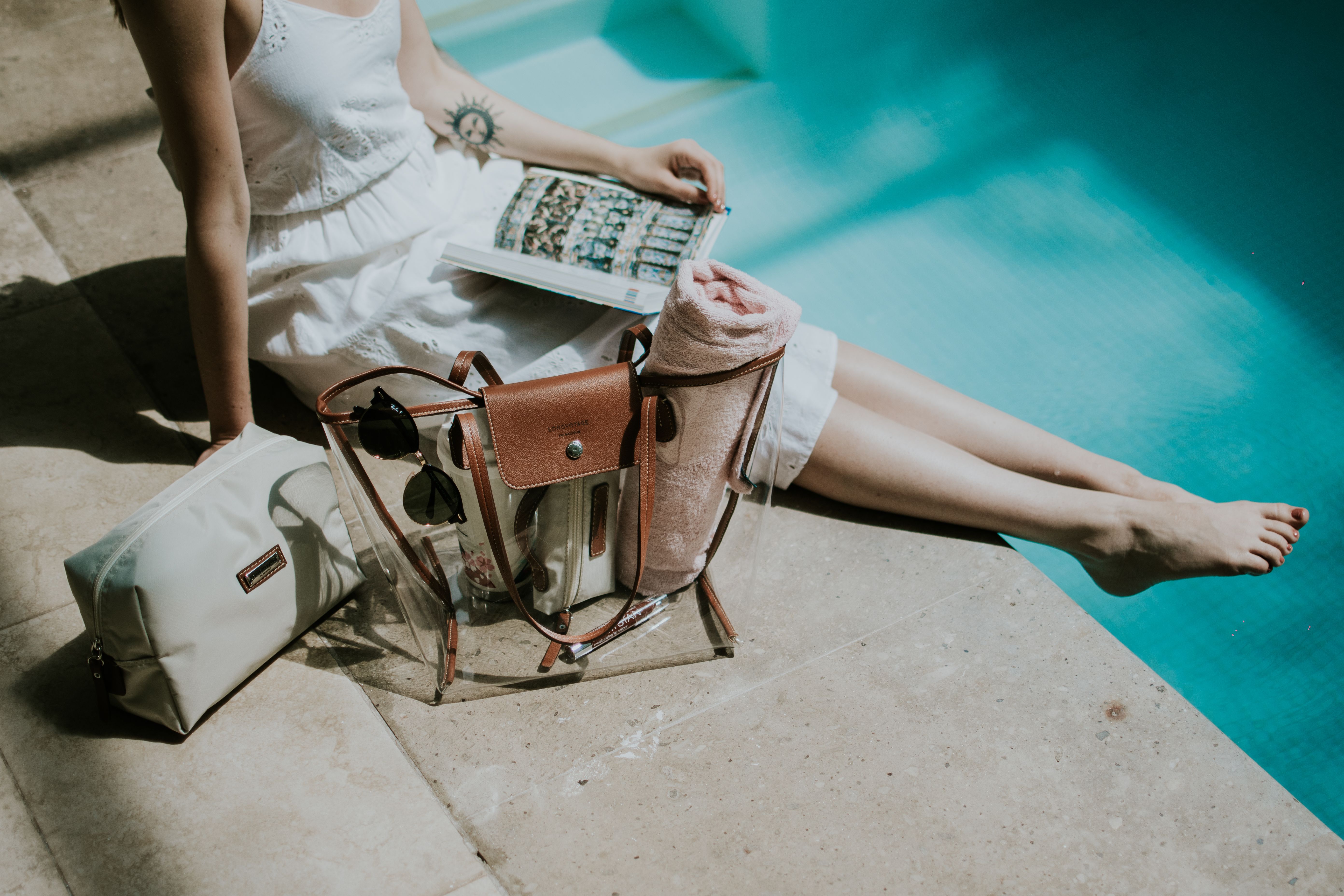 Woman enjoying a sunny day by the pool, reading a book with a transparent bag beside her.