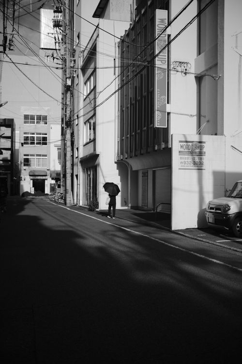Free A solitary person walks with an umbrella in a quiet black and white urban street, casting shadows. Stock Photo