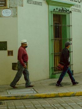 Two men walking on a sidewalk past a historic wall in Oaxaca, Mexico, showcasing cultural street life.