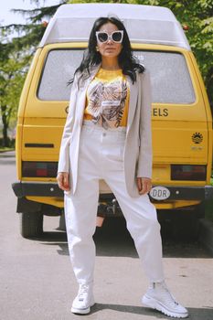 Confident woman in white outfit posing in front of yellow vehicle on a sunny day.